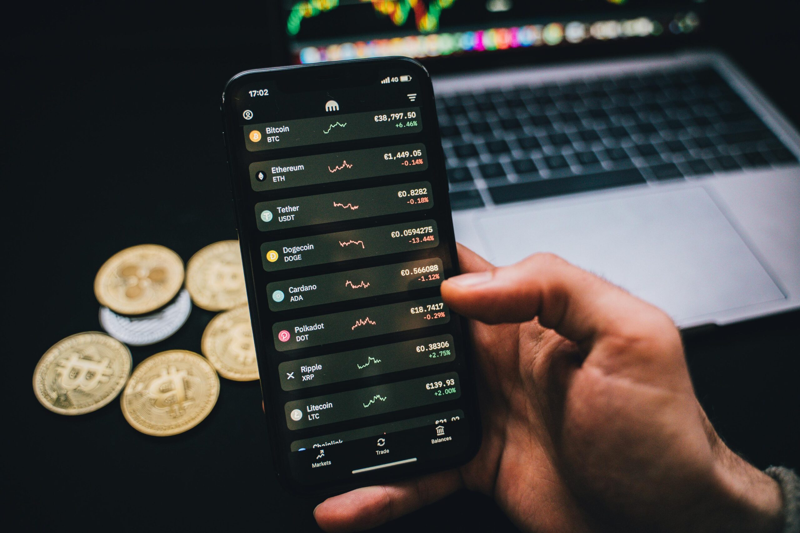 Home A person checks cryptocurrency values on a smartphone with a laptop and Bitcoins nearby.